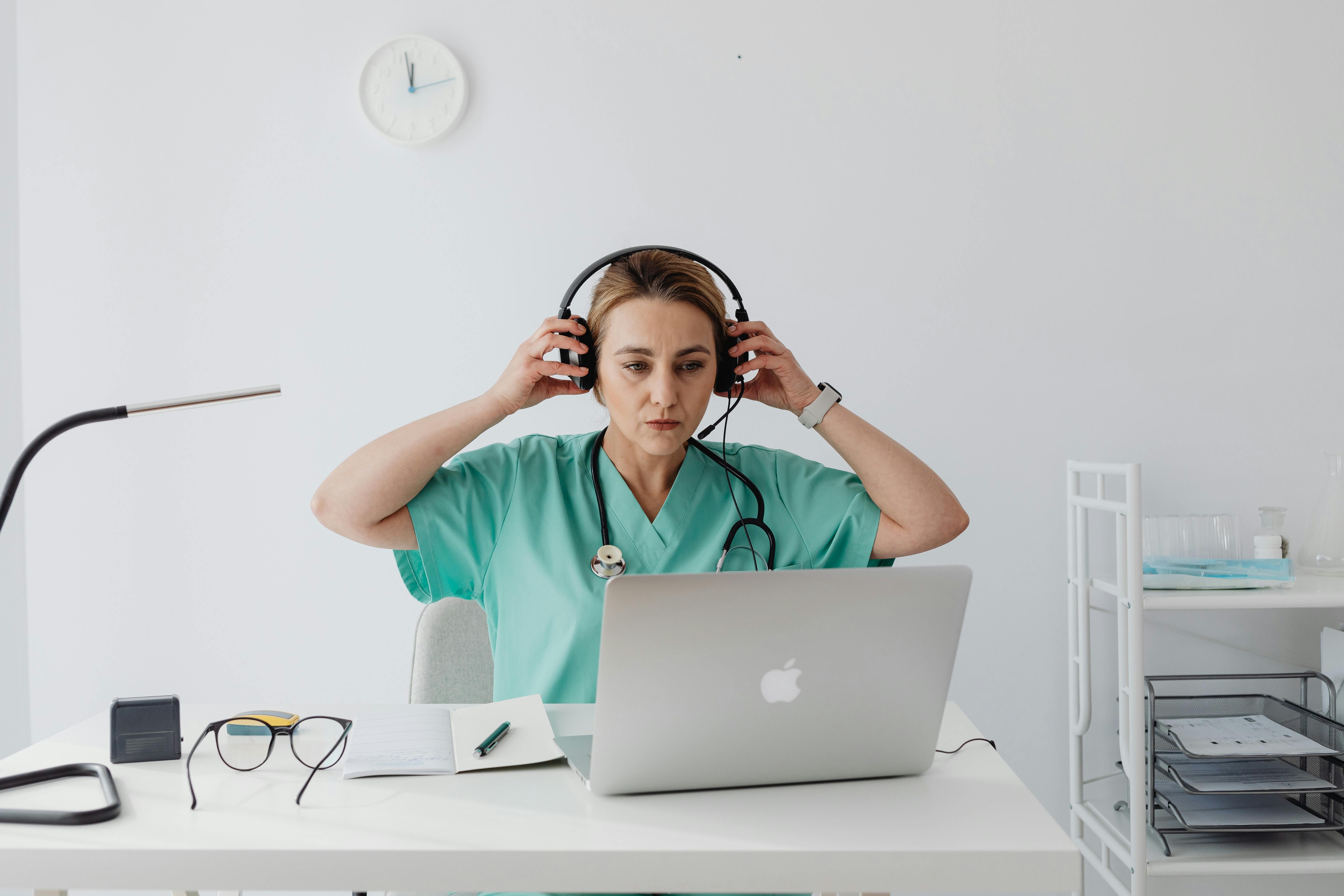 Healthcare worker in scrubs attending an online meeting or educational session with headphones and a laptop.