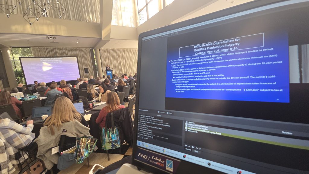 A view from behind a laptop running broadcasting software, displaying a blue presentation slide titled "100% Elective Depreciation for Qualified Production Property." In the background, an audience sits at round tables in a conference hall, facing a speaker at a podium and a large projection screen showing the same presentation.