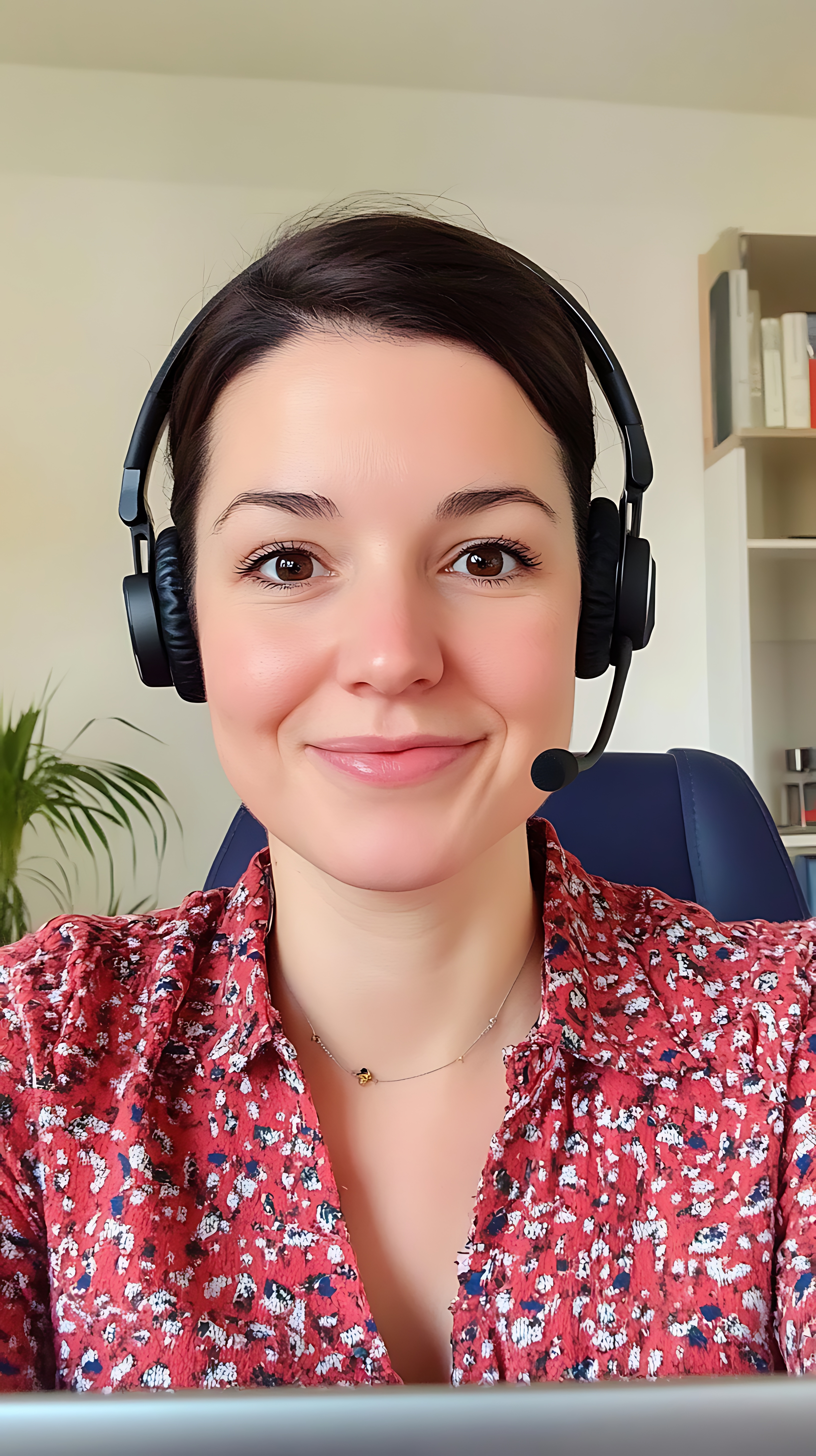A person wearing a dark blue shirt with white polka dots is sitting in front of a laptop. The person is wearing a headset with a microphone. The background shows a cozy room with a bookshelf, a lamp, and a window with curtains