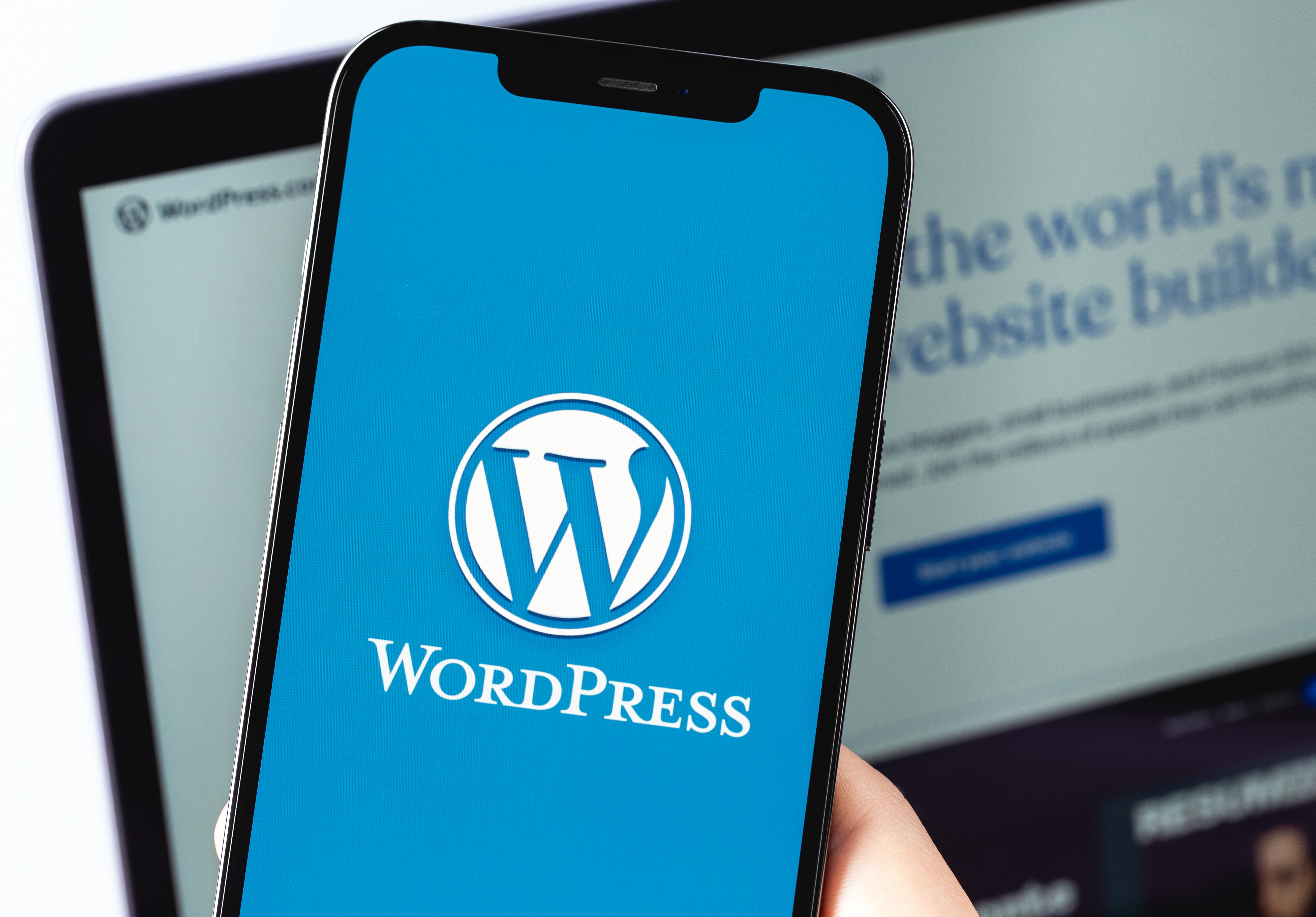 A close-up of a hand holding a smartphone that prominently displays the official WordPress logo on a blue screen. In the background, a blurred laptop screen is visible showing a website interface.