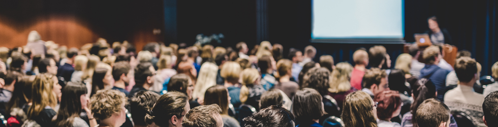 Crowded conference hall with attendees facing a stage and a presenter at a podium near a screen.